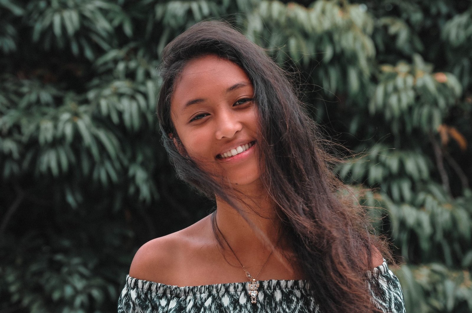 A young woman with flowing hair smiles outdoors with lush green foliage background.